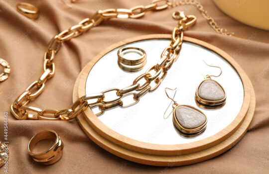 Stylish golden bijouterie and mirror on light brown fabric, closeup